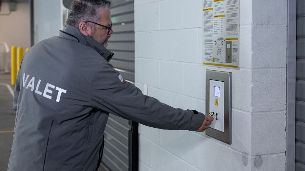 Imaginea prezintă o persoană la panoul de control al unui sistem automatizat de parcare, care confirmă procesul de stocare și astfel inițiază transportul automat al vehiculului către un loc de parcare disponibil.
