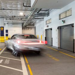 Imaginea prezintă un vehicul care intră într-un sistem de parcare automatizat dintr-o parcare subterană modernă, cu mai multe porți de acces.